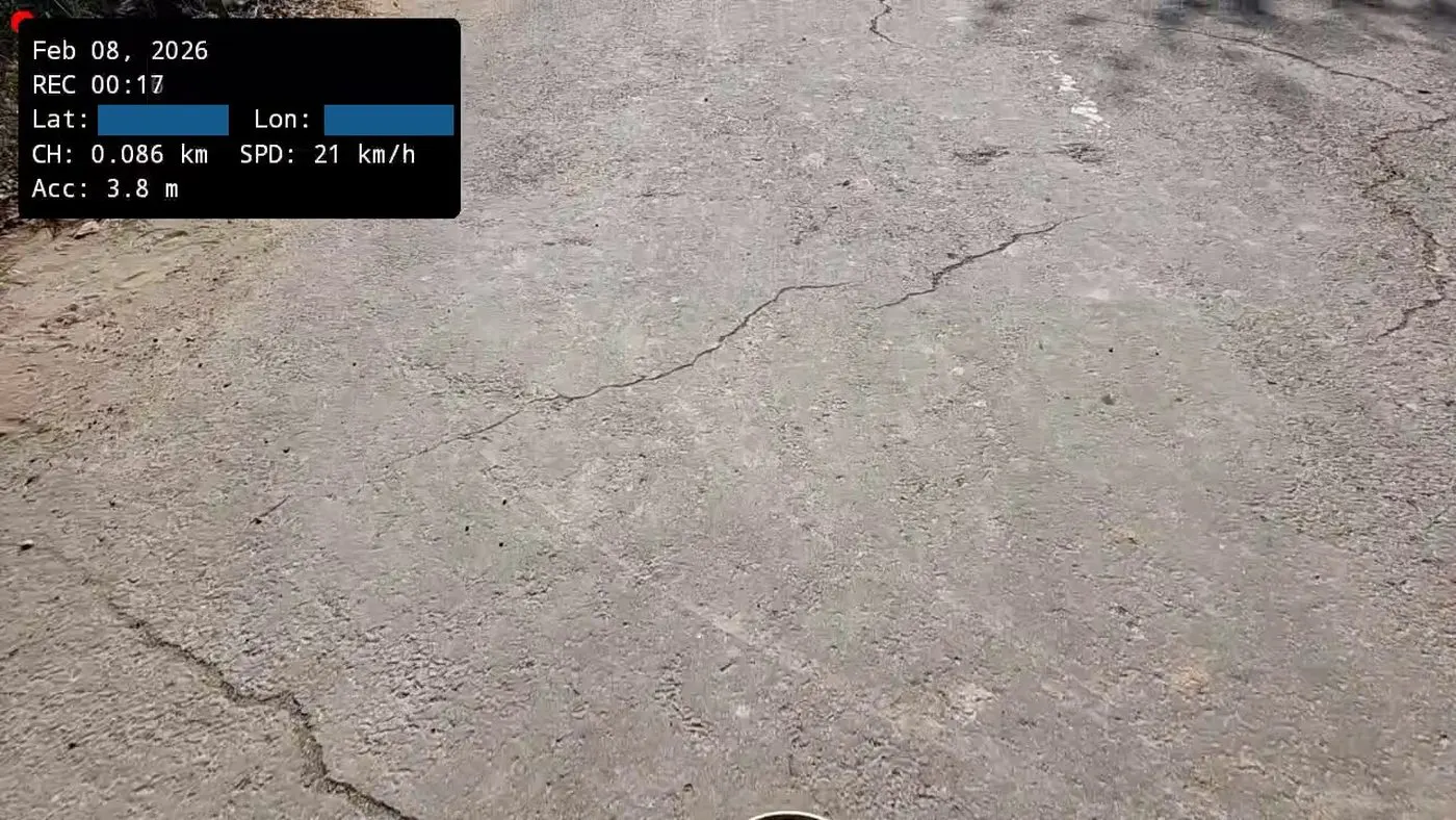 A frame recorded by the Chainage Android app. A panel burnt into the top-left corner shows the date, recording time, chainage of 0.086 km, speed of 21 km/h and GPS accuracy of 3.8 m. Latitude and longitude are covered by solid bars. Below it, a cracked concrete road surface fills the frame.